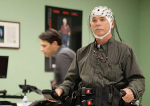 A man wearing a cap of neural or brain sensors stands with the assistance of a robotic exoskeleton device.