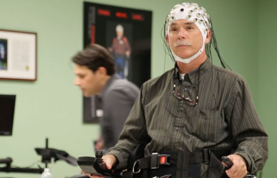 A man wearing a cap of neural or brain sensors stands with the assistance of a robotic exoskeleton device.